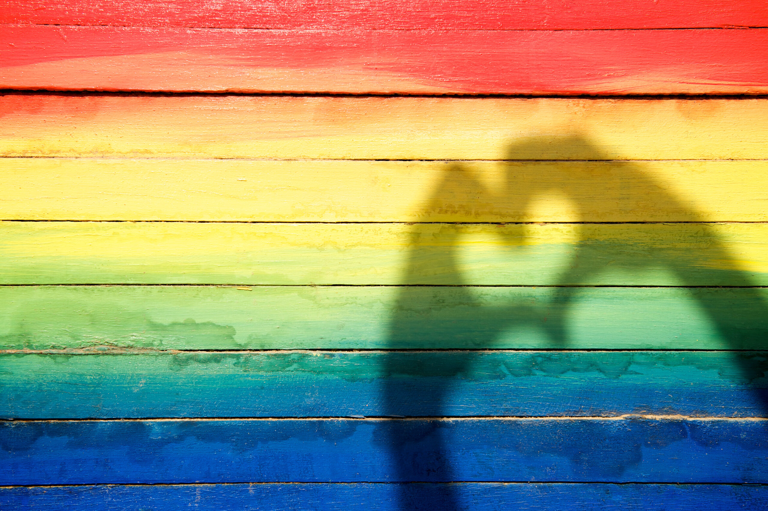 Skygge av hender som former et hjerte, på en regnbuebakgrunn.