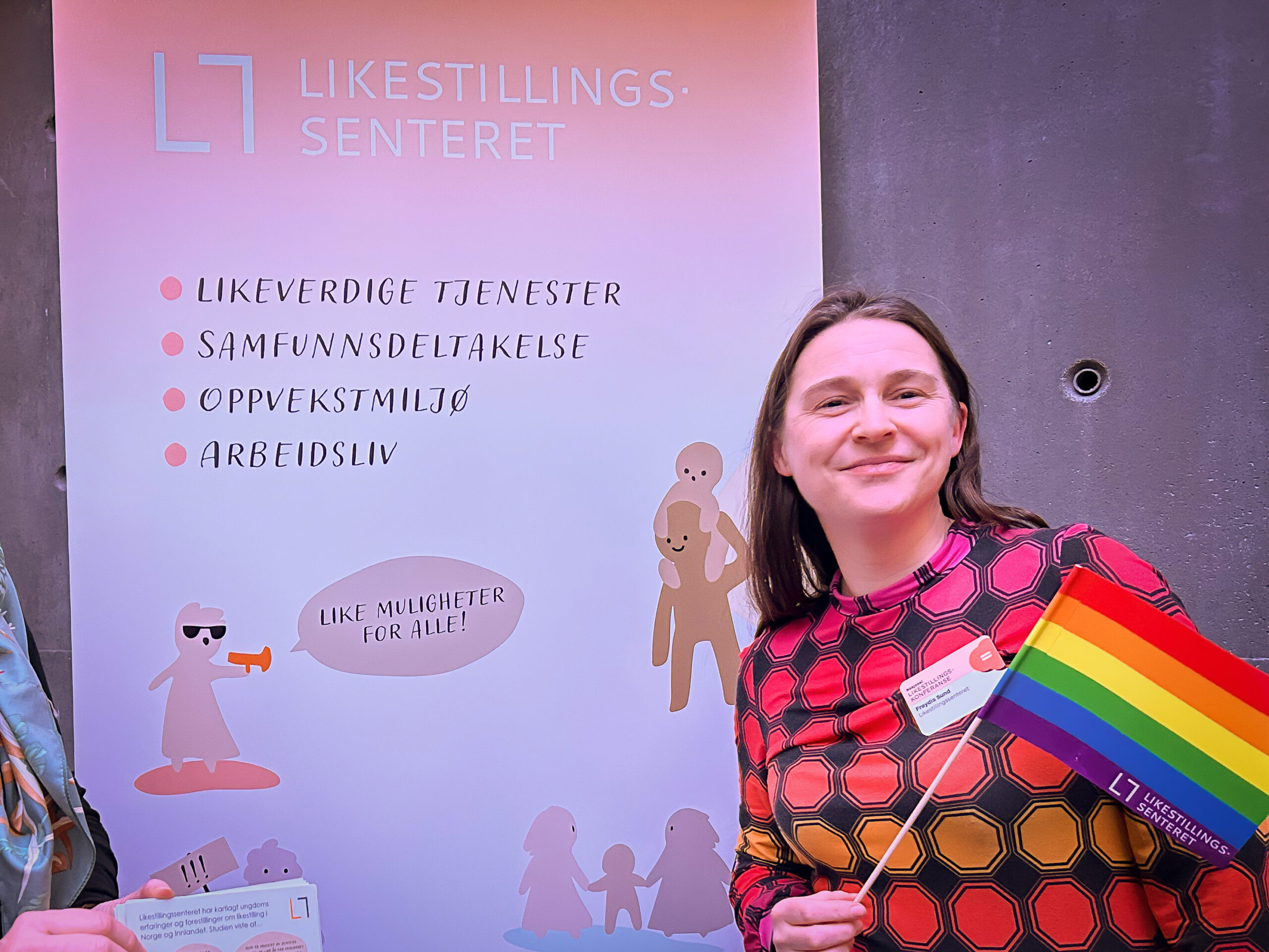Frøydis Sund står foran en rollup med Likestillingssenterets logo. Hun smiler og holder et pride-flagg.