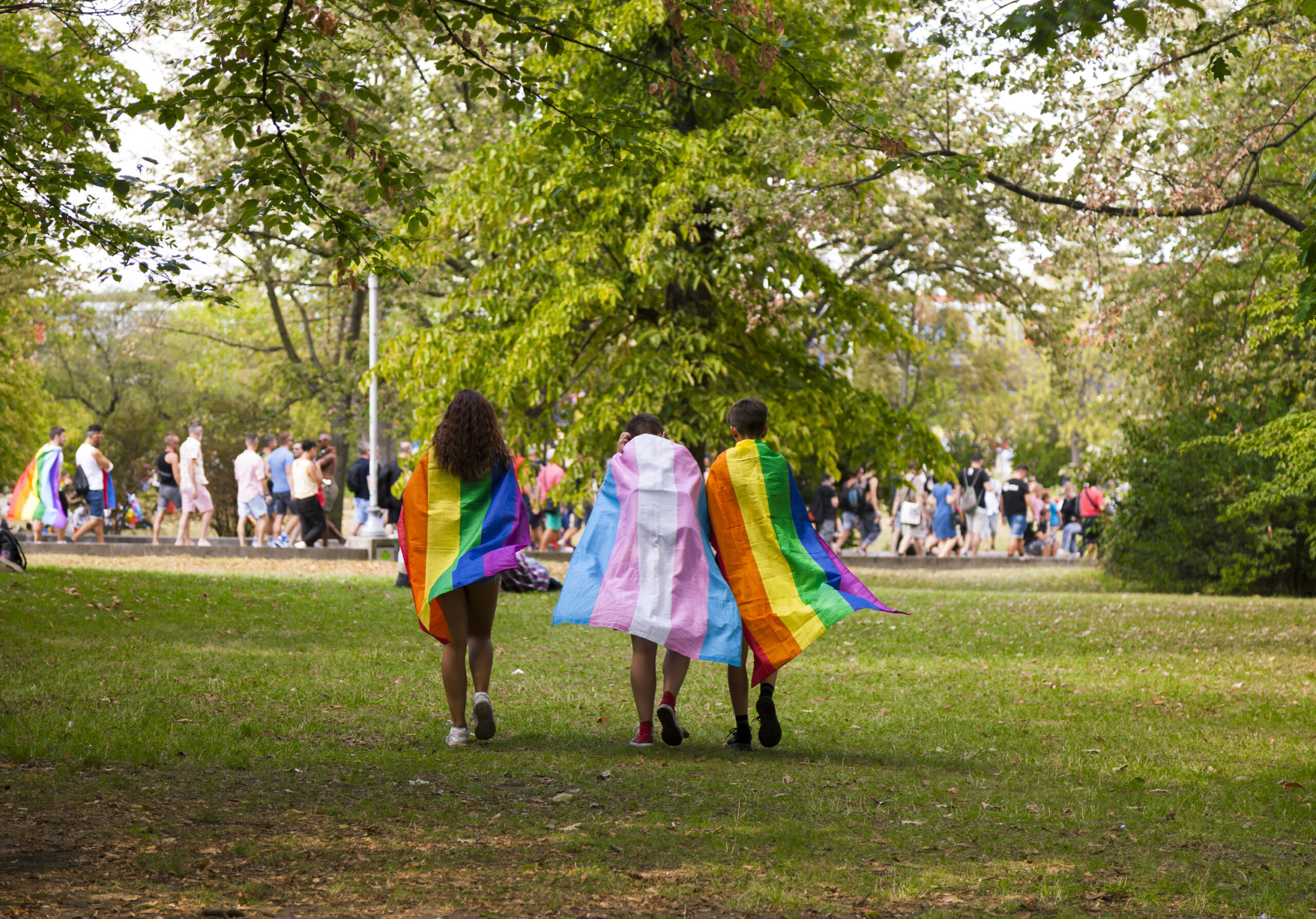 Tre personer går gjennom en park. To av dem har regnbueflagg som kappe, den siste har flagget for transpersoner.