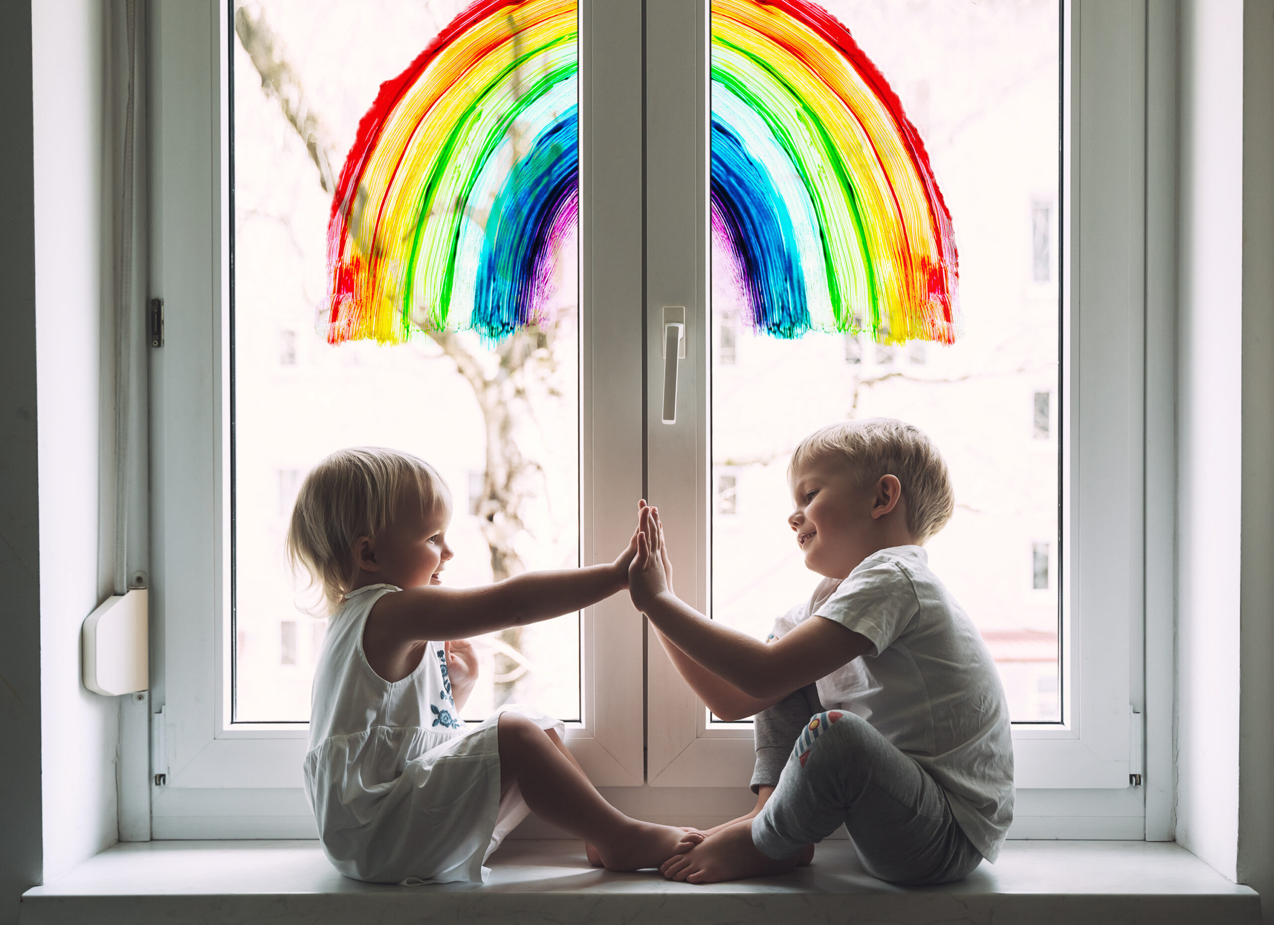 To barn sitter og leker i en vinduskarm. På vinduet er det malt en regnbue.