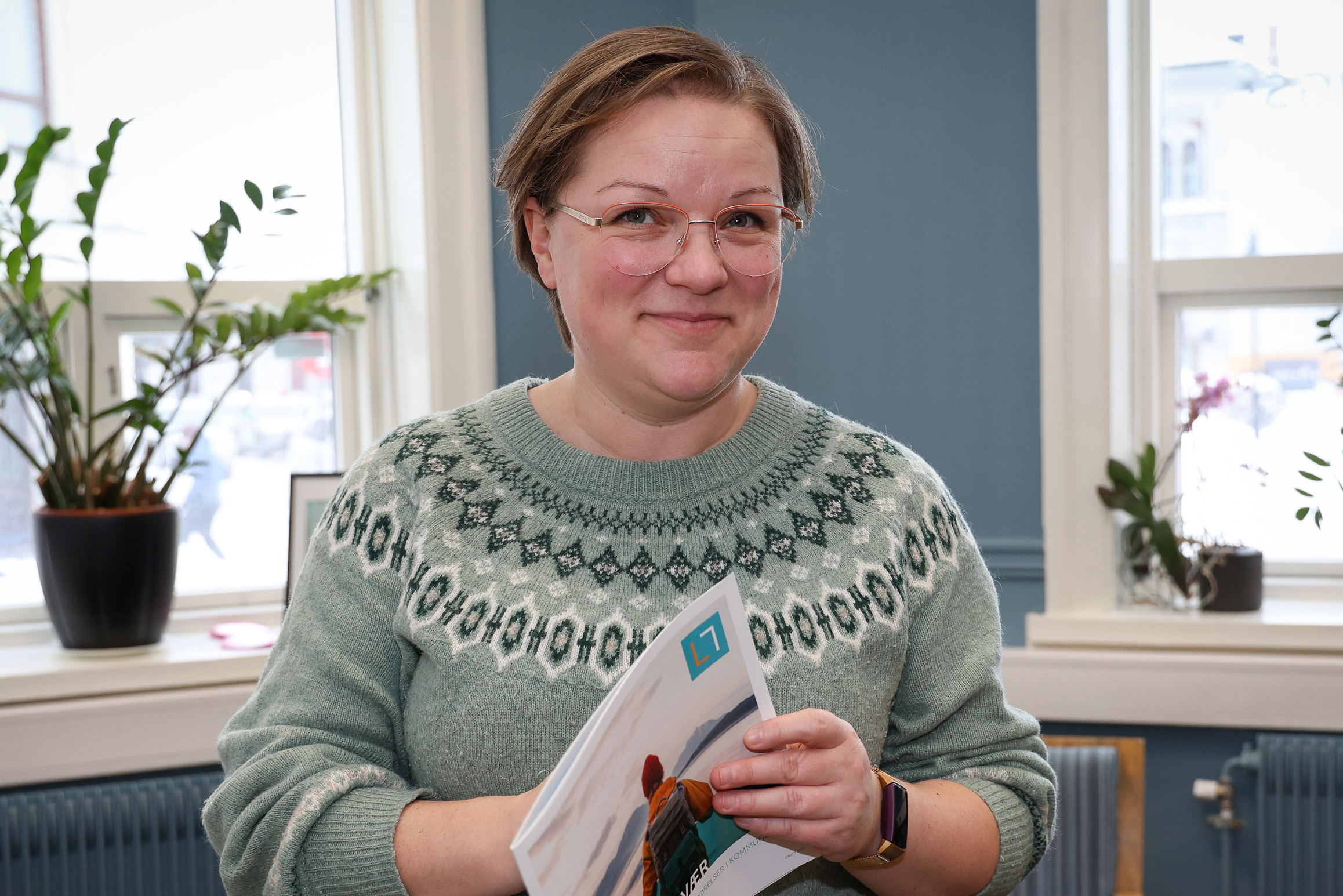 Annette Solberg står og smiler til kamera, holder en brosjyre.