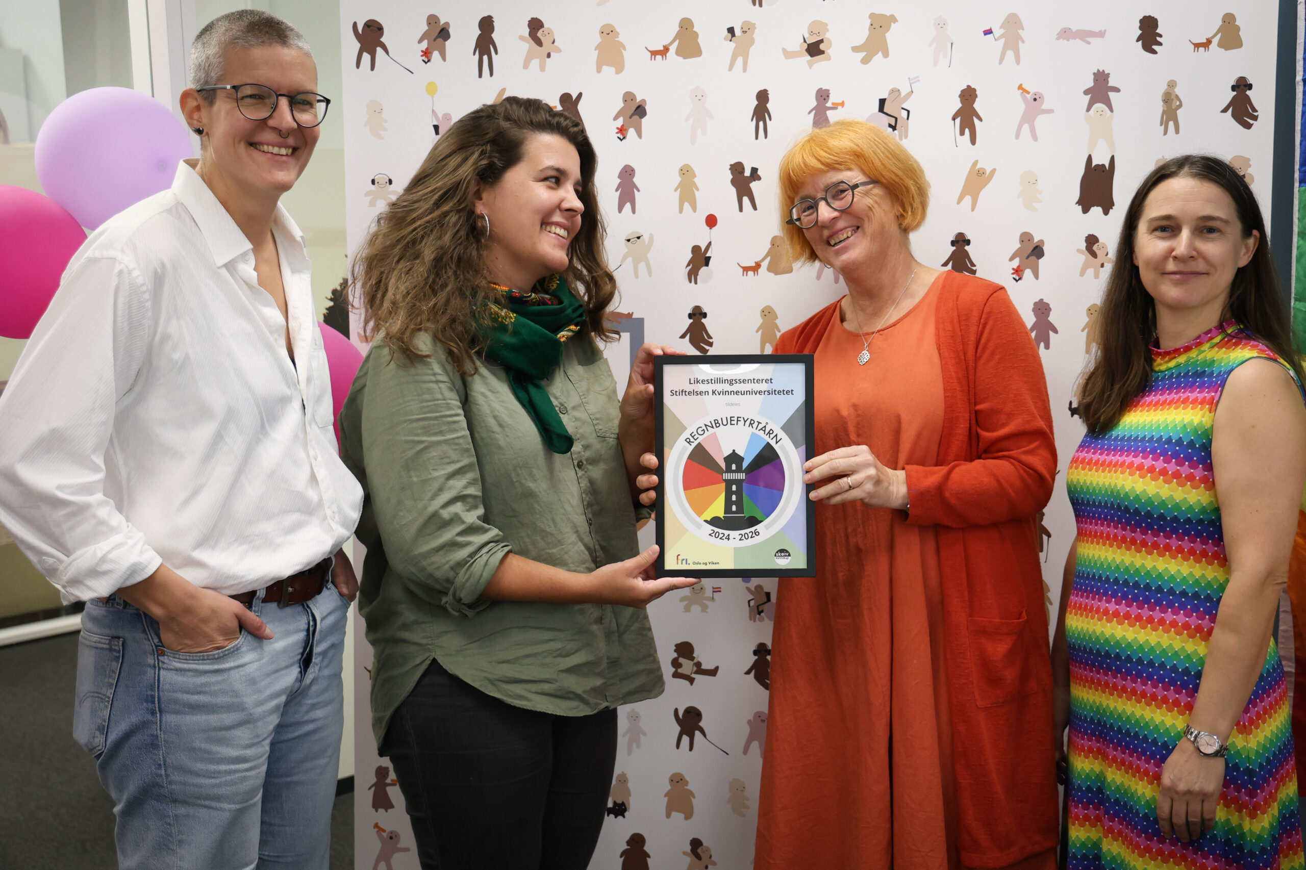 Fire personer står på rekke, den ene får overrakt et Regnbuefyrtårn-diplom i en ramme. De smiler og det er god stemning.