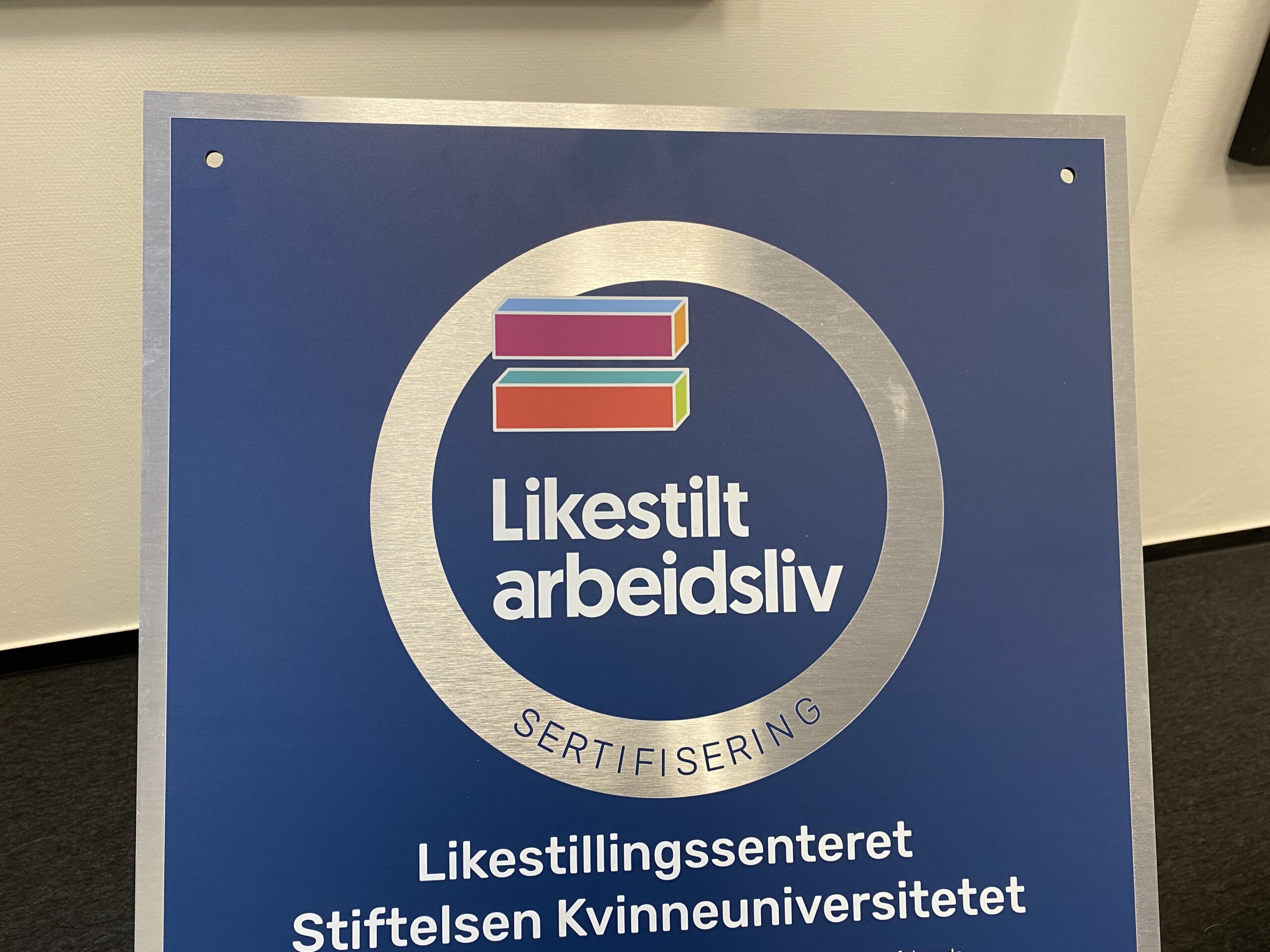 Logo til Likestilt Arbeidsliv står på mørkeblå bakgrunn. Et fargerikt er lik-tegn er plassert over teksten.