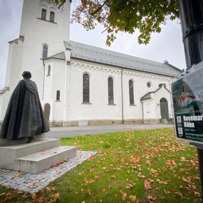 I bildet ser vi en plakett med info om biskop Rosemarie Köhn og hennes statue. I bakgrunnen er Hamar domkirke.