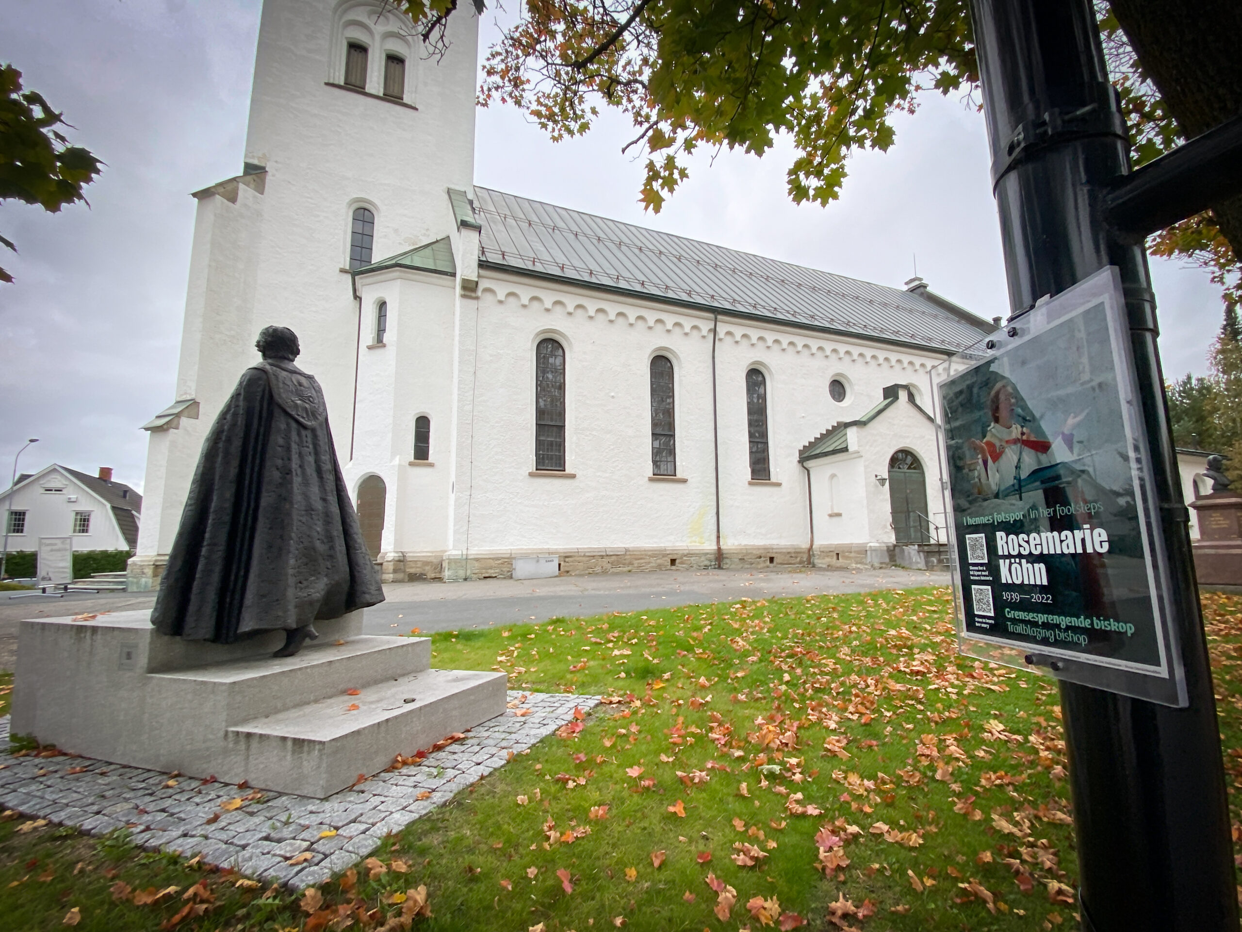 I bildet ser vi en plakett med info om biskop Rosemarie Köhn og hennes statue. I bakgrunnen er Hamar domkirke.