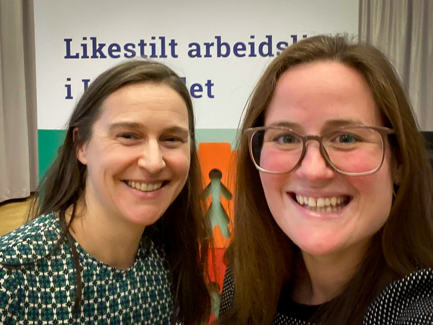Frøydis Sund og Kristina Miklavi og ser i kamerea og smiler. Bak dem er en rollup hvor teksten Likestilt arbeidsliv i Innlandet er synlig.