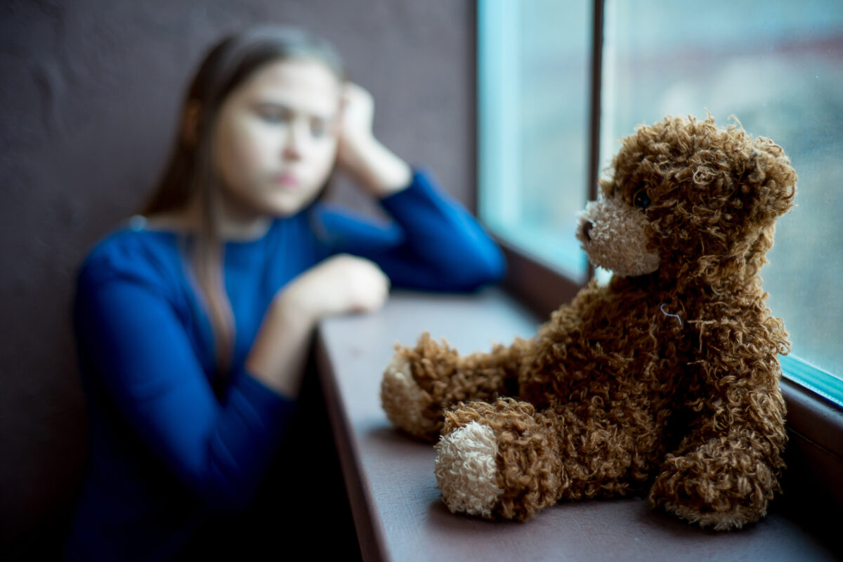 
			I forgrunnen sitter en bamse lent mot et vindu. Uskarpt i bakgrunnen sitter et barn og lener hodet på hånda.			