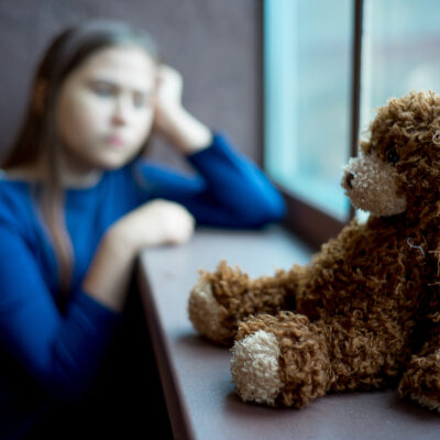 I forgrunnen sitter en bamse lent mot et vindu. Uskarpt i bakgrunnen sitter et barn og lener hodet på hånda.