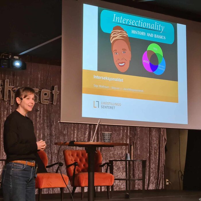 Silje Mathisen står på en scene. Bak henne vises en presentasjon hvor det står intersectionality.