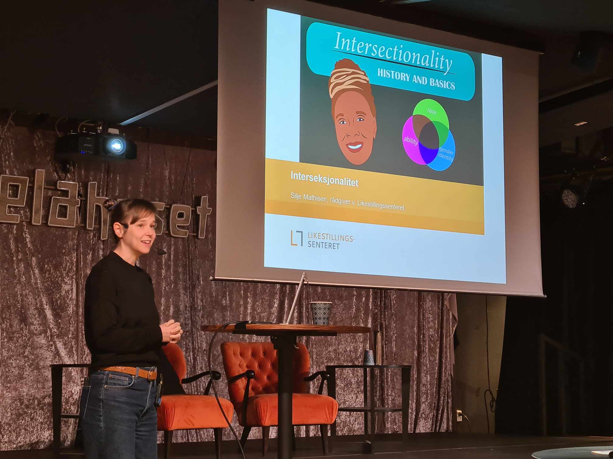 Silje Mathisen står på en scene. Bak henne vises en presentasjon hvor det står intersectionality.