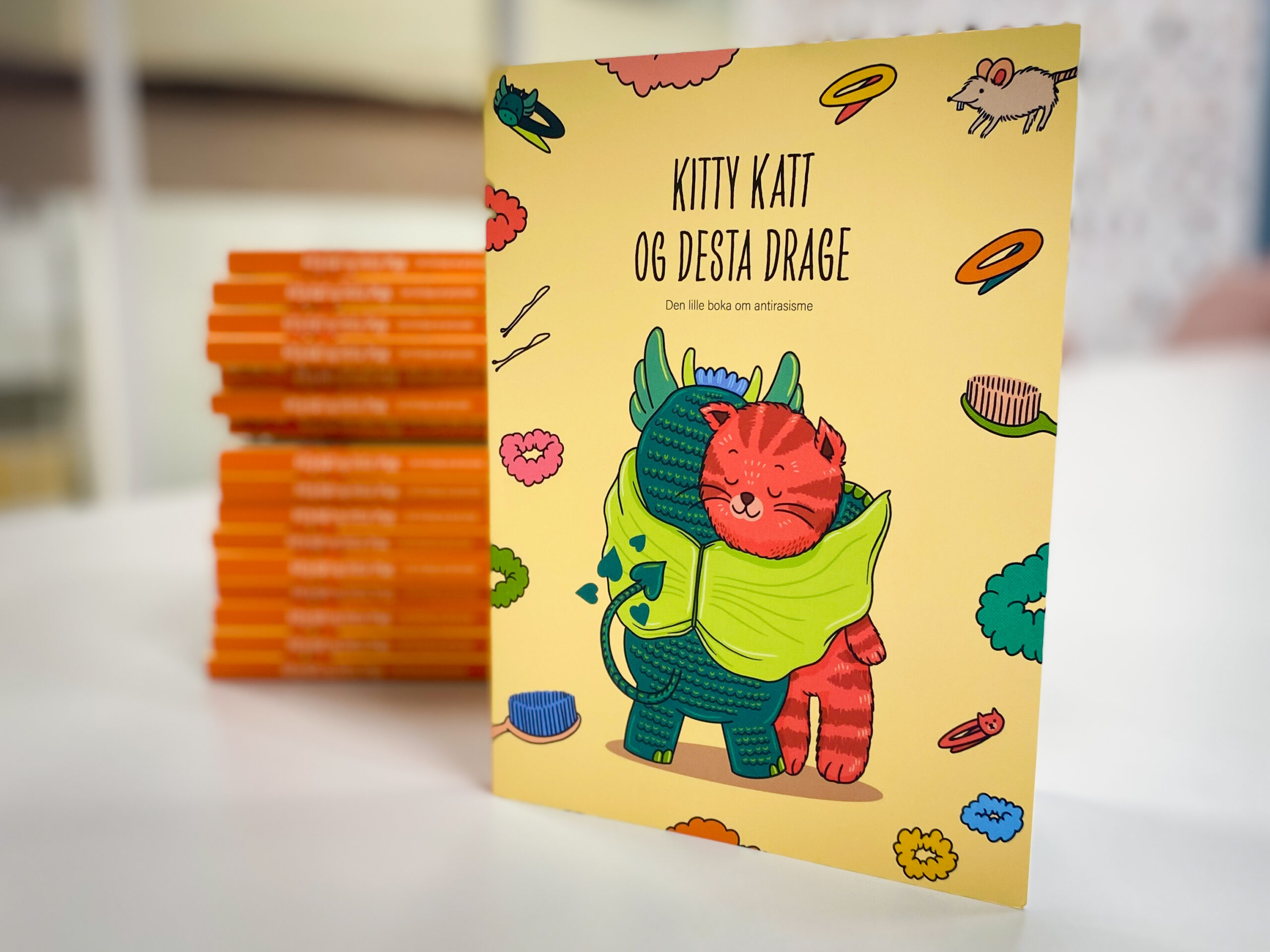 Bilde av omslaget til boka Kitty Katt og Desta Drage - den lille boka om antirasisme. En rød katt og en grønn drage klemmer hverandre.