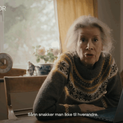 Stillbilde fra en film. En eldre dame sitter foran en PC, hun ser i kamera. Under står teksten "sånn snakker man ikke til hverandre".