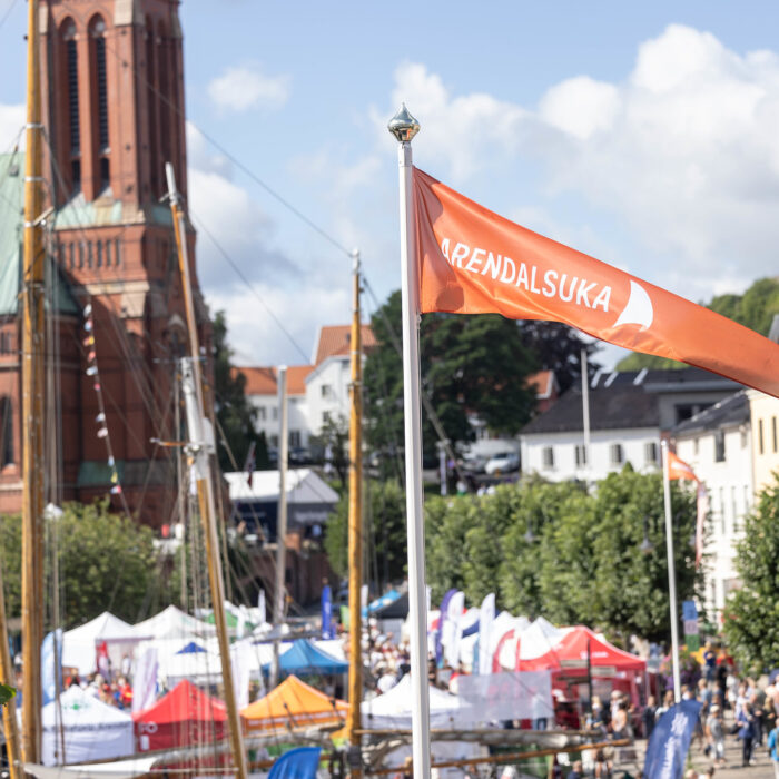 Et oransje banner med teksten Arendalsuka vaier i vinden med byen i bakgrunn.