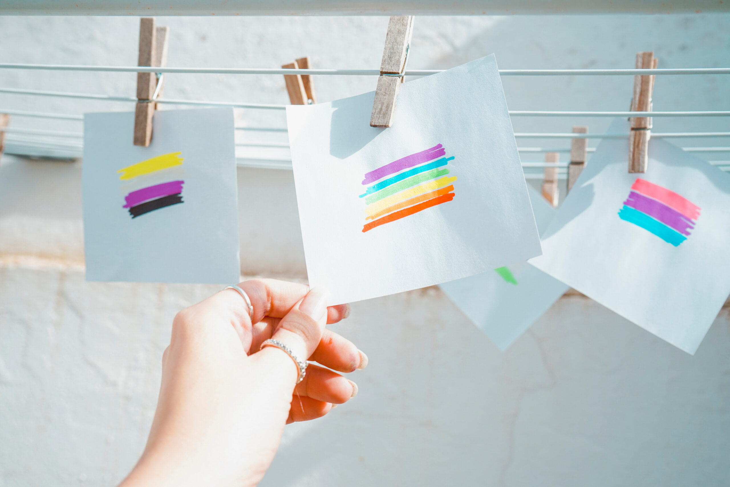 Et regnbueflagg er tegnet på et ark som henger på en tørkesnor. En hånd griper arket. Det henger flere ark med ulike flagg i bakgrunnen.