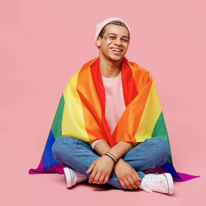 Full body smiling happy cheerful young gay man wear sweatshirt hat wrapped in flag sitting on floor isolated on plain pastel light pink color background studio portrait. Lifestyle lgbtq pride concept.