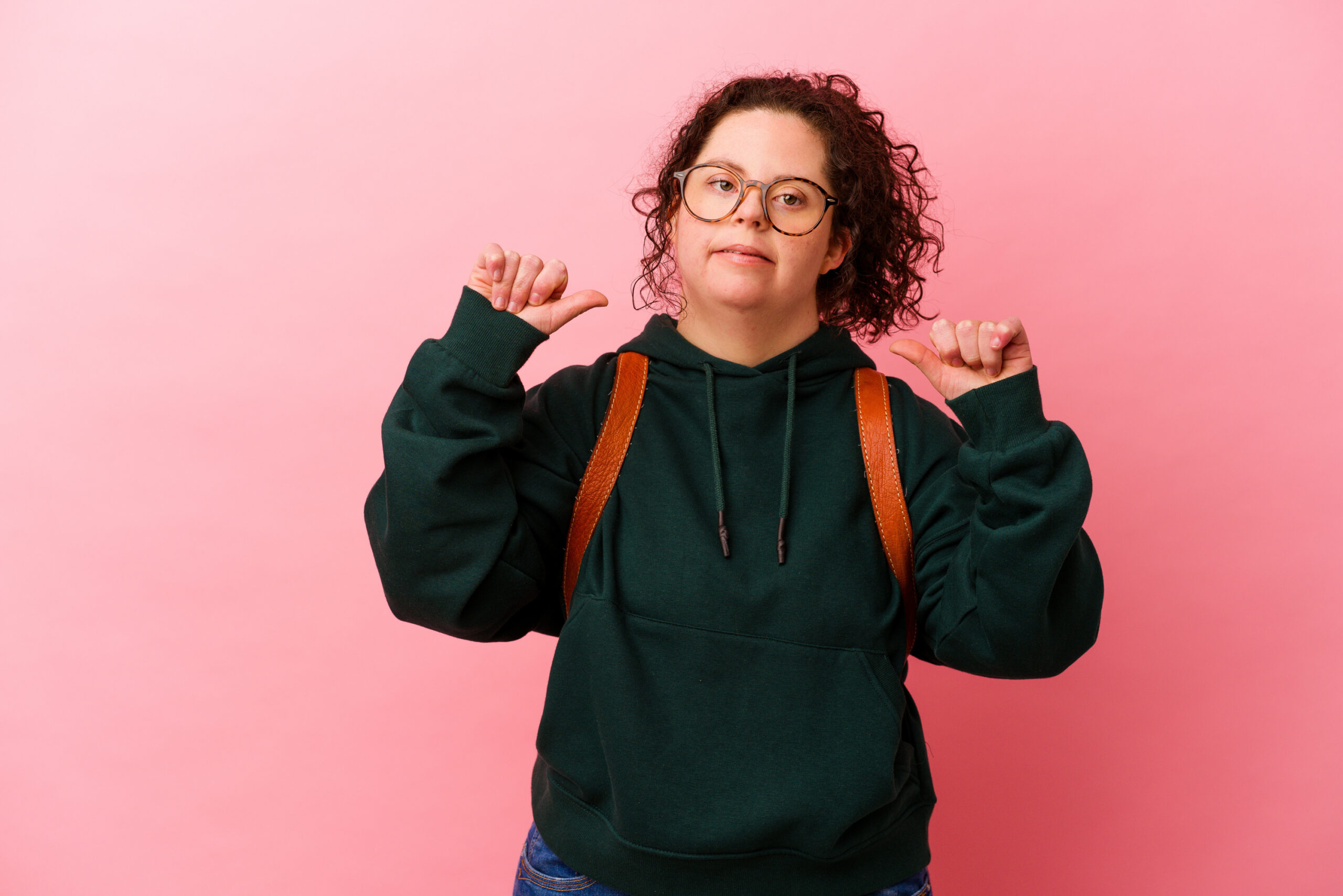 En person gir to tomler opp. Hun har ryggsekk, et lite smil og står foran en rosa bakgrunn.