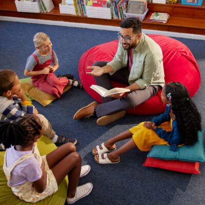 Barnehagelærer sitter sammen med fire elever og leser en bok.